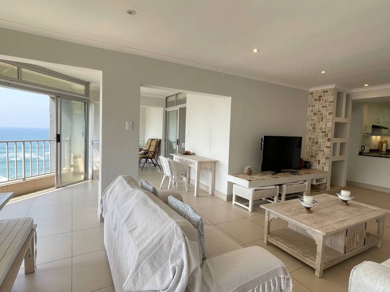 Spacious open-plan living area featuring tiled floors, recessed lighting, and a stone-clad feature wall, flowing seamlessly into a functional kitchen and balcony with ocean views.