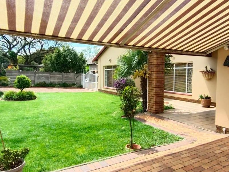 A covered patio area featuring a retractable striped awning, brick pillar support, and paved flooring, overlooking a well-maintained garden and driveway.