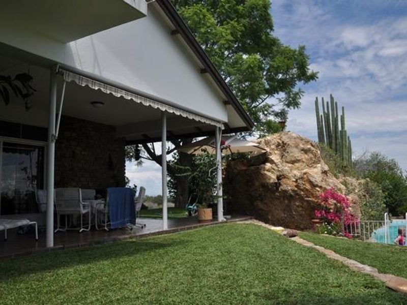 A covered patio area featuring a stone wall and retractable awning, overlooking a landscaped garden with a natural rock feature and swimming pool.