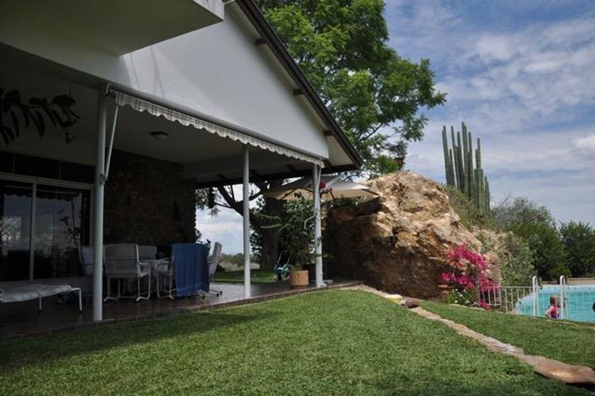 A covered patio area featuring a stone wall and retractable awning, overlooking a landscaped garden with a natural rock feature and swimming pool.
