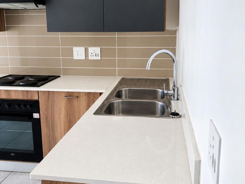 Modern kitchen featuring sleek dark upper cabinets, wood-grain lower cabinetry, stone countertops, integrated oven, hob, and a double stainless steel sink.