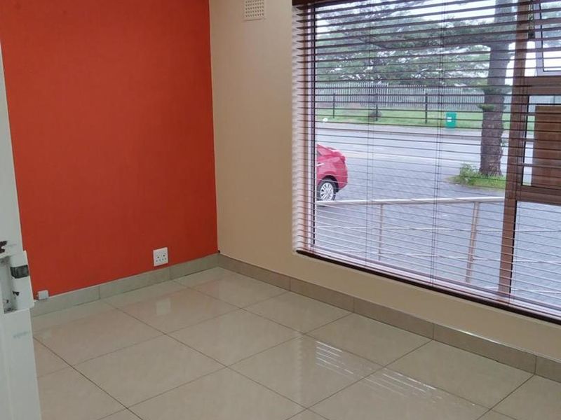 A bright, empty office space featuring tiled flooring, a split-system air conditioner, and a large window with venetian blinds overlooking the street.