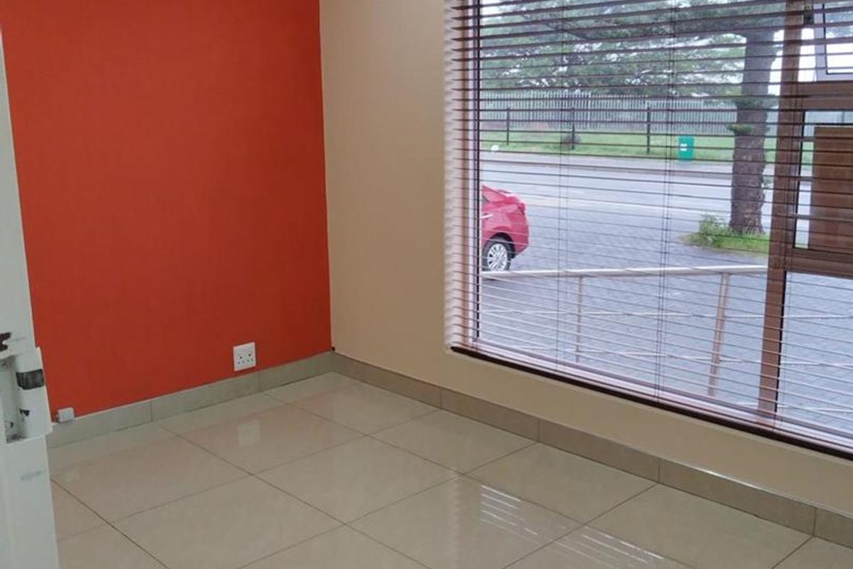 A bright, empty office space featuring tiled flooring, a split-system air conditioner, and a large window with venetian blinds overlooking the street.