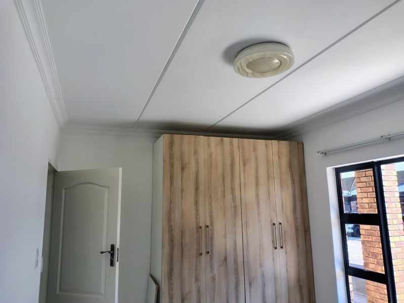 A clean, bright bedroom featuring modern wood-finish built-in cupboards, neutral walls, and a contemporary ceiling light fixture.
