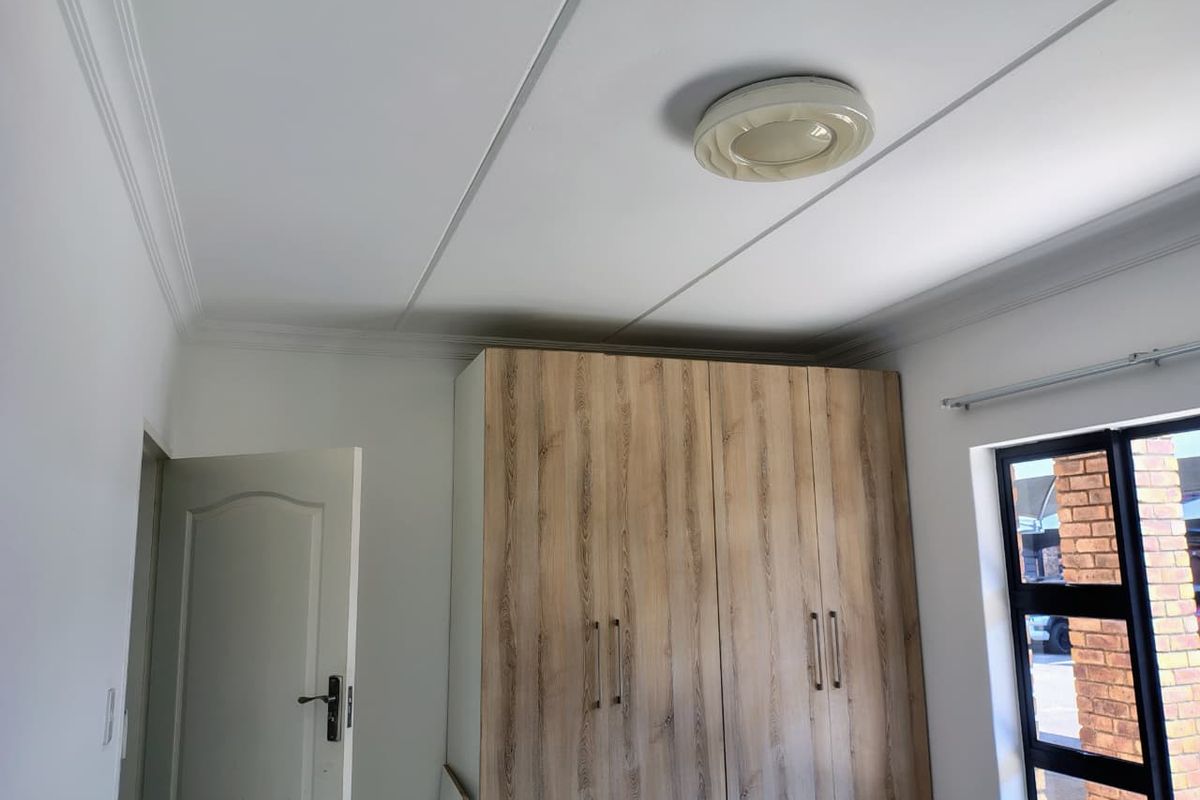 A clean, bright bedroom featuring modern wood-finish built-in cupboards, neutral walls, and a contemporary ceiling light fixture.
