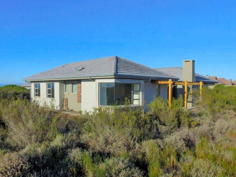 Rear view of a single-story house featuring a tiled roof, chimney, and timber pergola, surrounded by natural coastal vegetation.