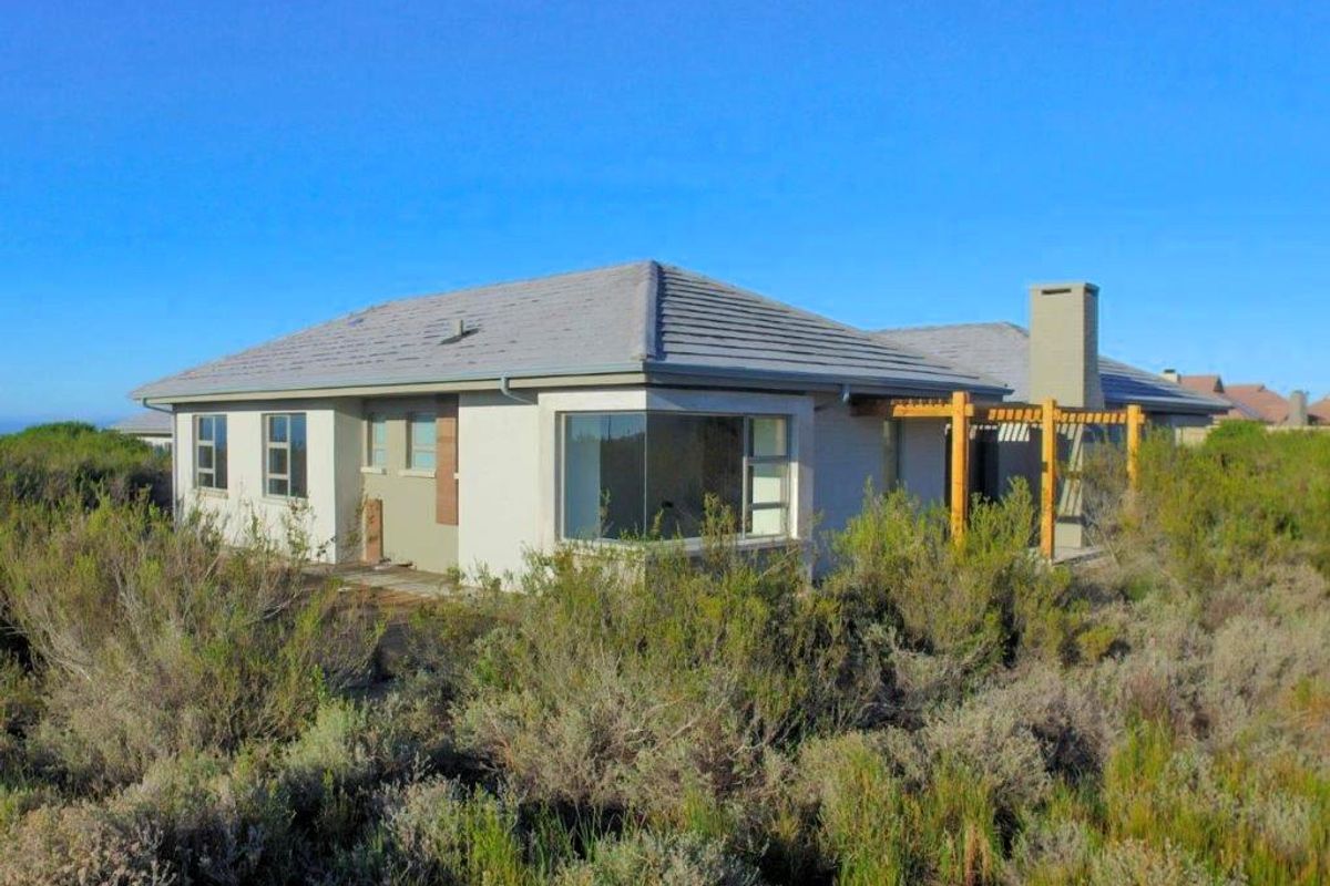 Rear view of a single-story house featuring a tiled roof, chimney, and timber pergola, surrounded by natural coastal vegetation.
