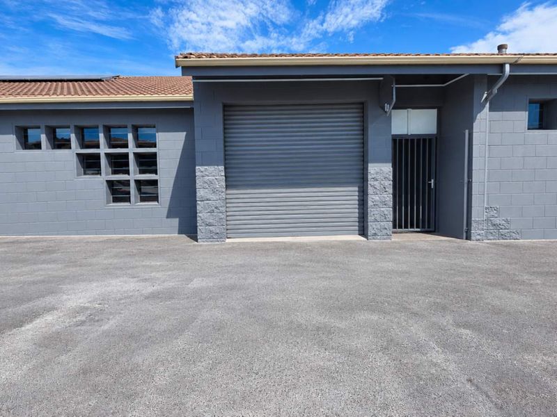 Industrial property exterior featuring grey concrete block walls, multiple roller shutter doors, and a secure entrance gate with an asphalt forecourt.