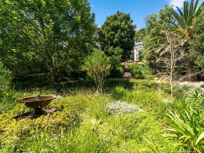 A lush, established garden featuring mature trees, diverse plantings, and a decorative stone fountain, with stone steps leading toward the rear of the residence.