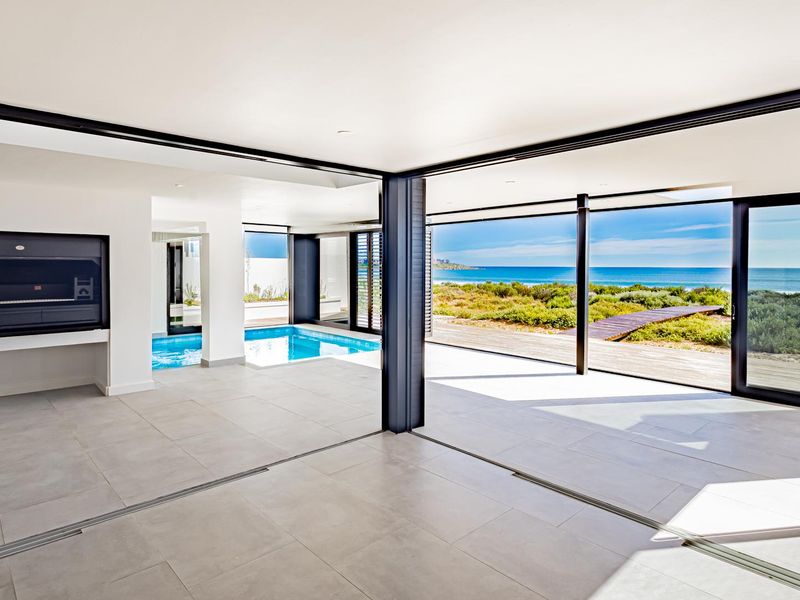 Spacious open-plan living area featuring large sliding glass doors, an indoor swimming pool, and a built-in braai with stunning coastal views.