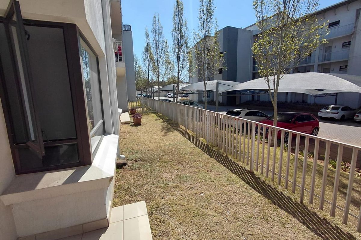 A ground floor apartment exterior featuring a small private lawn area, tiled patio section, and metal perimeter fencing overlooking the complex parking area.