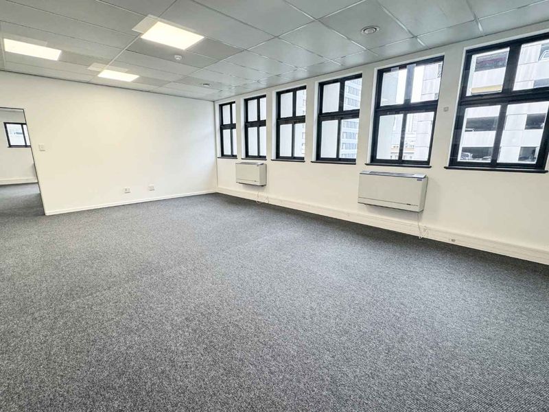 A bright, open-plan office space featuring neutral carpet flooring, a suspended ceiling with recessed lighting, and large windows with wall-mounted HVAC units.
