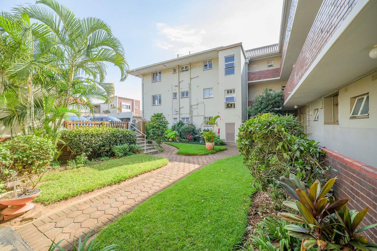 A view of the apartment complex exterior featuring a paved walkway, landscaped garden areas, and multi-story residential building facade.