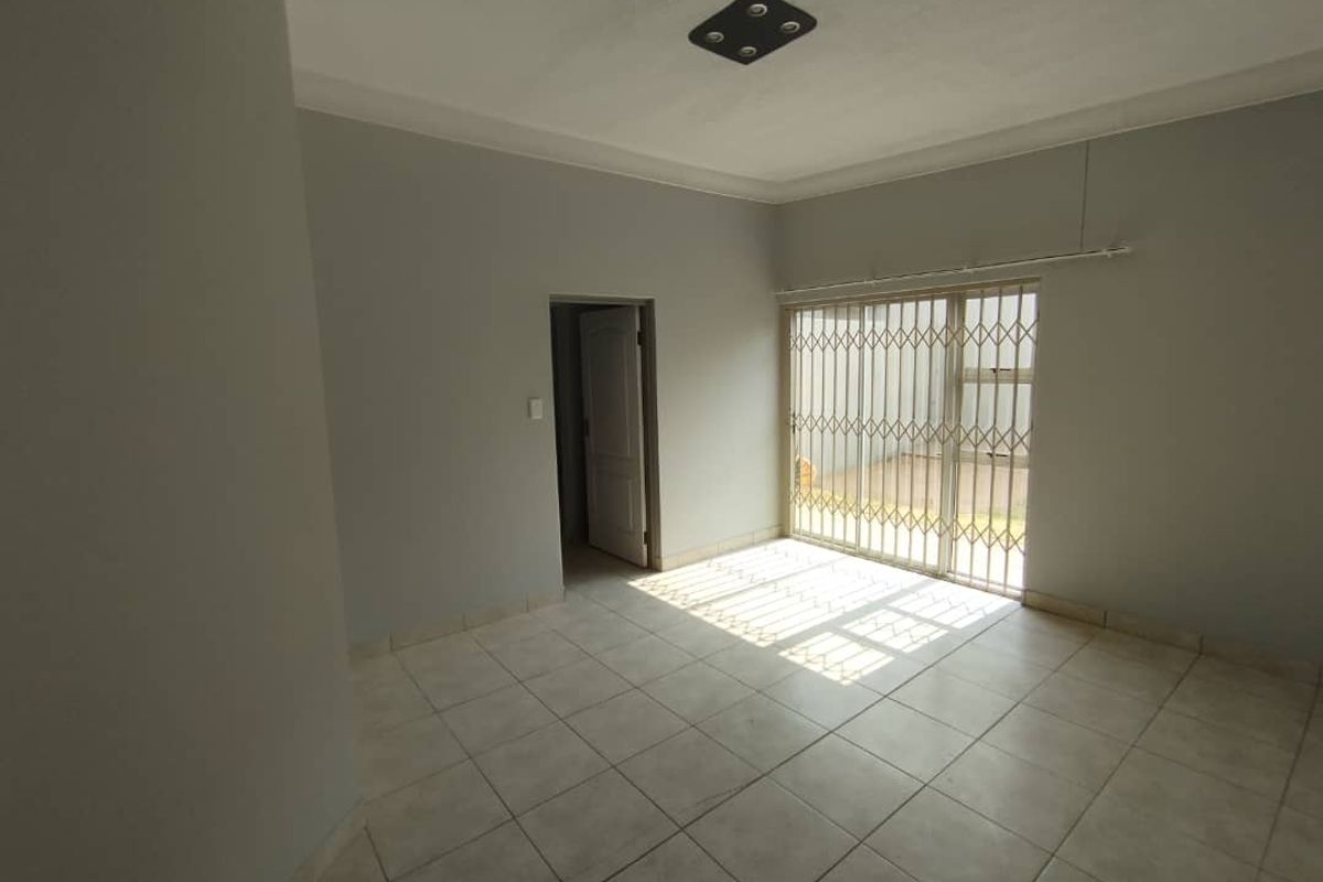 An empty living room featuring neutral tiled flooring, a ceiling light fixture, and a sliding glass door with a security gate leading outside.