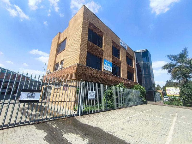 A three-story brick office building featuring a glass-enclosed stairwell, secure metal perimeter fencing, and a paved parking area.