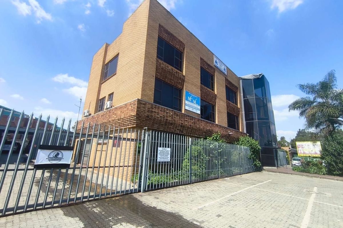 A three-story brick office building featuring a glass-enclosed stairwell, secure metal perimeter fencing, and a paved parking area.