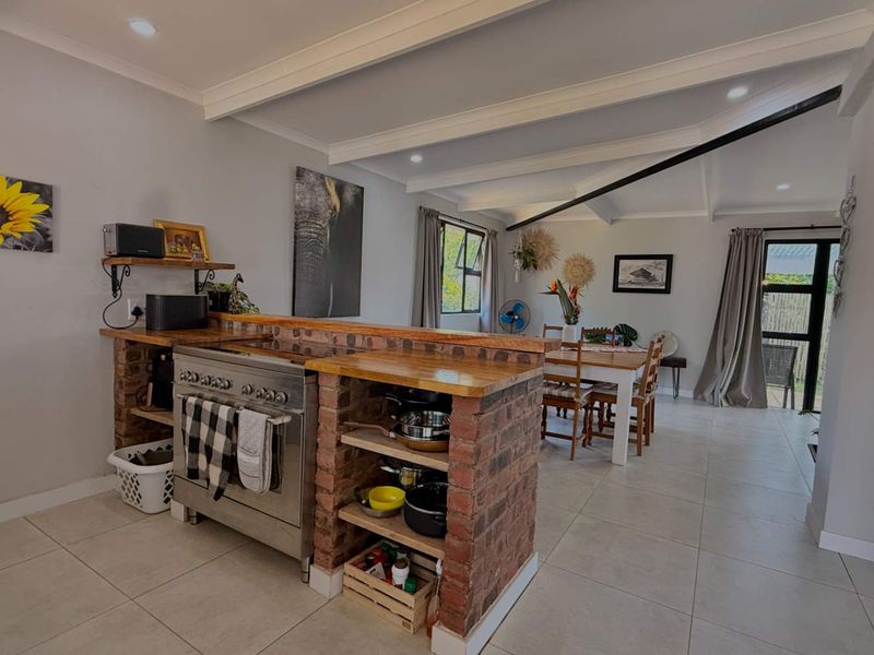 Open-plan living and dining area featuring a unique brick kitchen island with timber countertop, exposed beam ceilings, and neutral tiled flooring throughout.