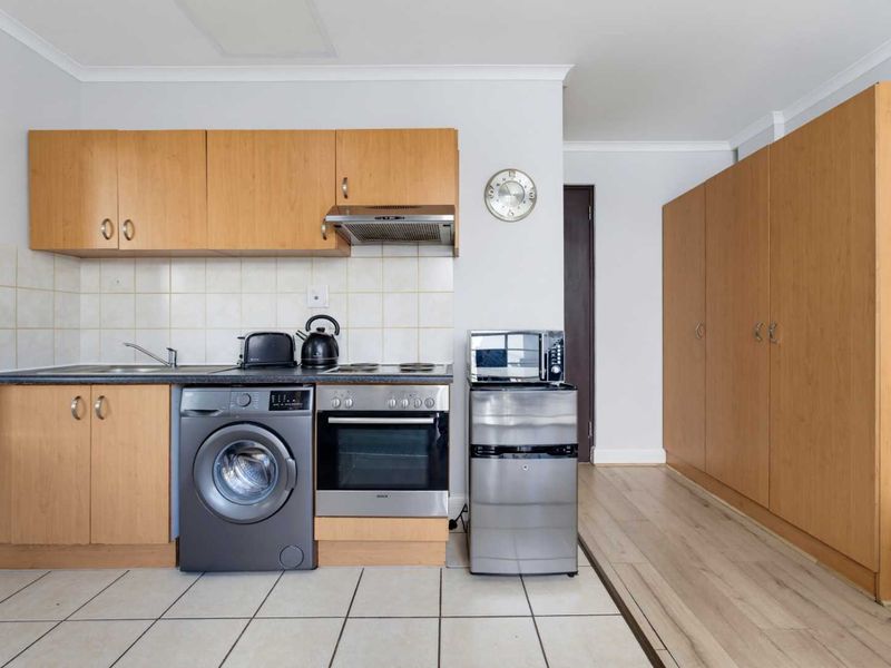A compact kitchen featuring light wood cabinetry, tiled backsplash, built-in oven and hob, and integrated laundry plumbing, adjacent to a storage cupboard area.