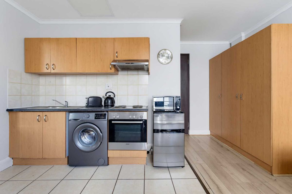 A compact kitchen featuring light wood cabinetry, tiled backsplash, built-in oven and hob, and integrated laundry plumbing, adjacent to a storage cupboard area.