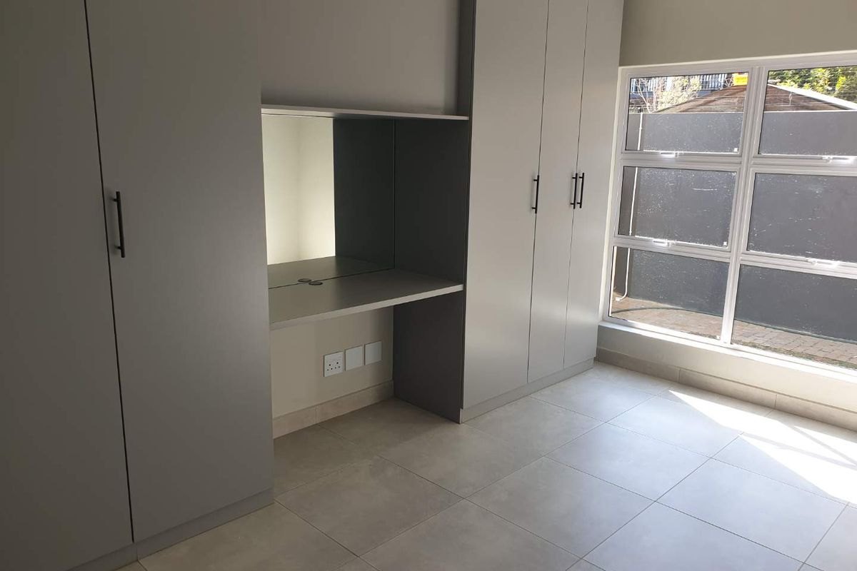 A modern bedroom featuring built-in grey cabinetry with an integrated desk and mirror, complemented by large windows and neutral floor tiling.