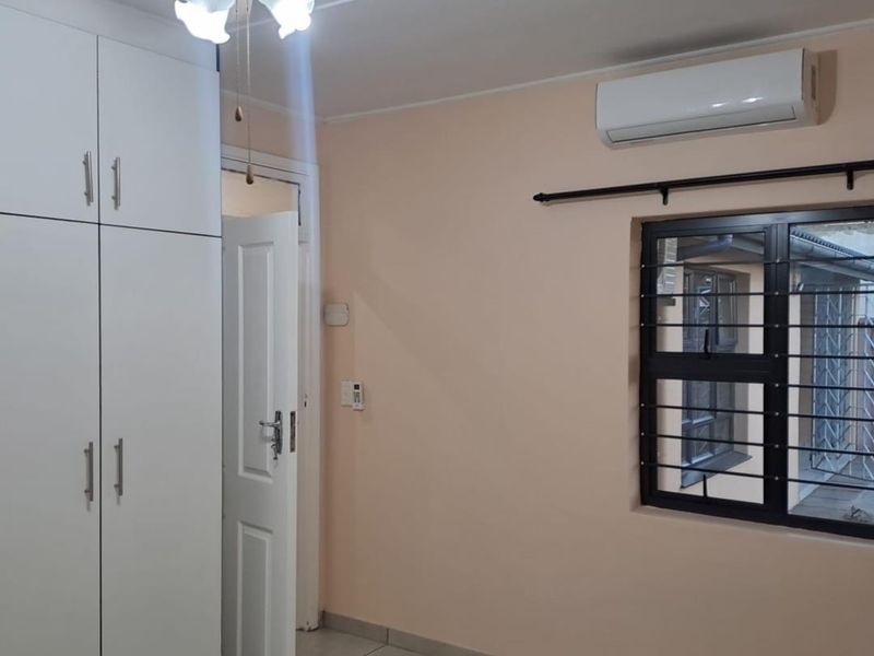 A clean, empty bedroom featuring built-in cupboards, a ceiling fan, split-system air conditioning, and tiled flooring with security bars on the window.