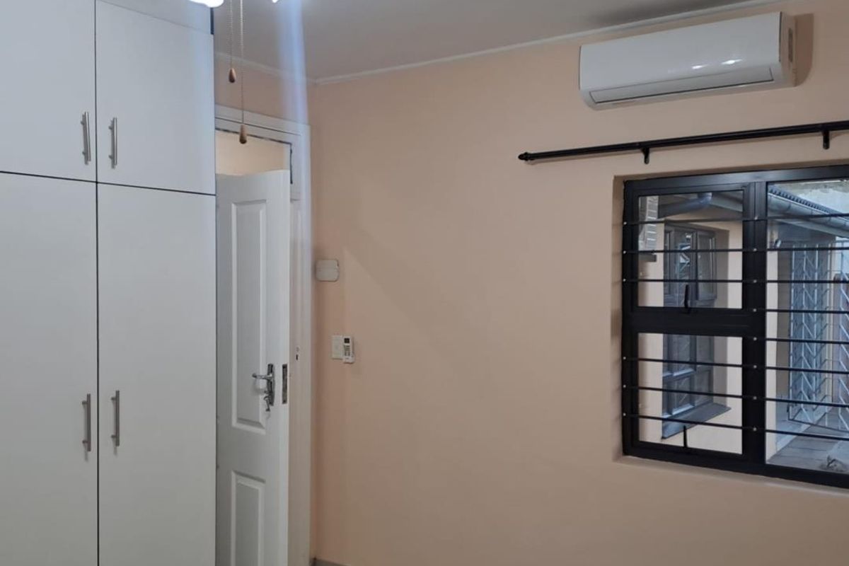 A clean, empty bedroom featuring built-in cupboards, a ceiling fan, split-system air conditioning, and tiled flooring with security bars on the window.