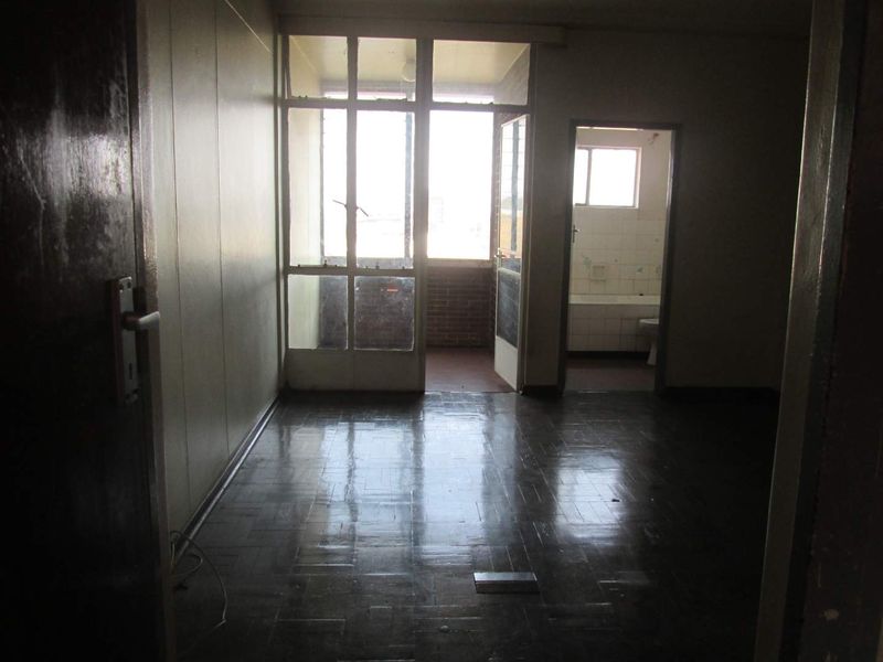 An empty living room featuring dark parquet flooring and access to an adjoining balcony and bathroom, requiring cosmetic updates to modernize the space.