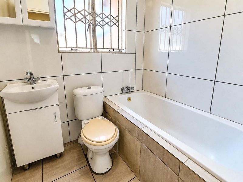 A functional bathroom featuring a vanity basin, toilet, and bathtub with full-height wall tiling and a security-fitted window.