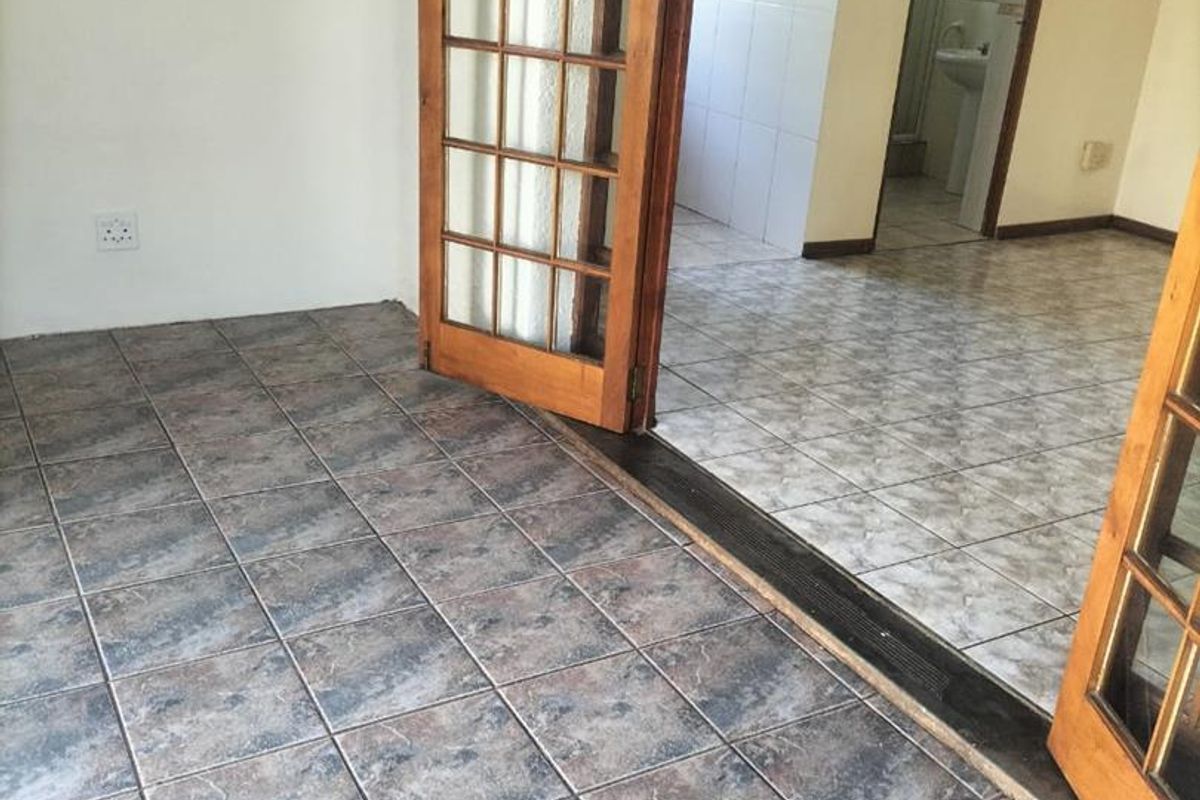 An empty living area featuring tiled flooring and wooden french doors leading to an adjacent room with a bathroom visible.