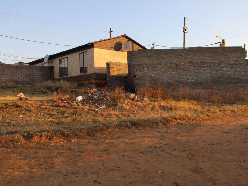 Exterior view of a residential property featuring a brick structure, tiled roof, and surrounding boundary walls on an unpaved plot.