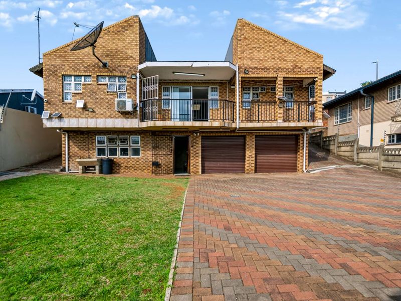 Rear view of a double-story face brick house featuring a paved driveway, double garage, and an upper-level balcony with metal railings.