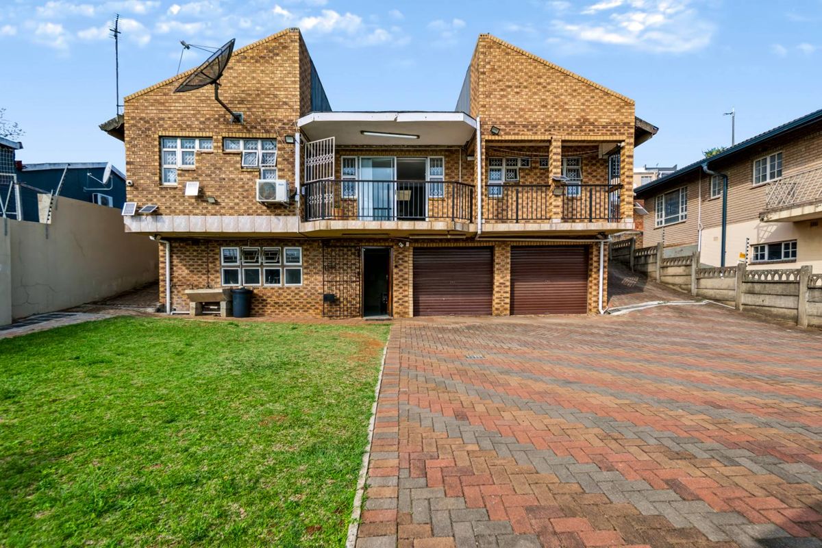 Rear view of a double-story face brick house featuring a paved driveway, double garage, and an upper-level balcony with metal railings.