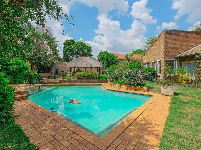 A well-maintained outdoor pool area featuring a swimming pool, brick paving, and a traditional thatched lapa set within a landscaped garden.