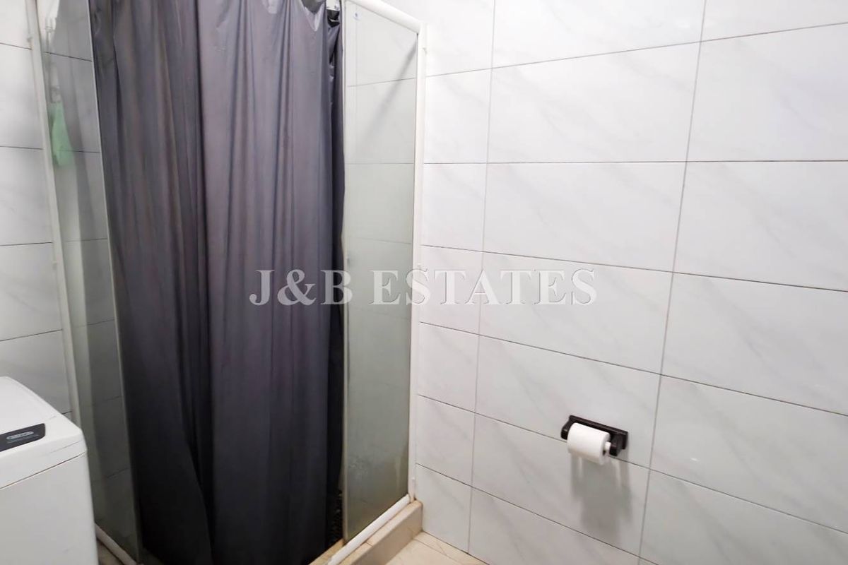 A functional bathroom featuring white wall tiles, a shower enclosure with a curtain, and a wall-mounted toilet paper holder.