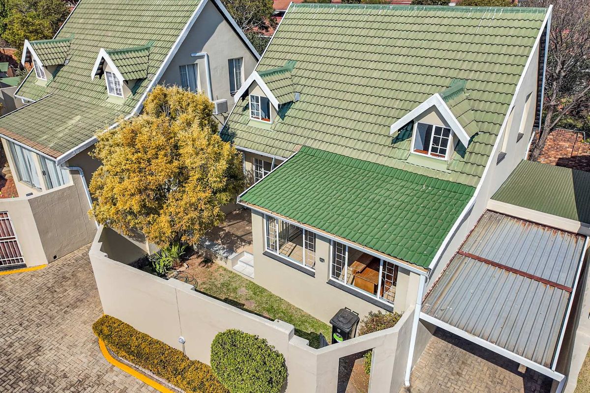 Aerial view of a townhouse unit featuring a green tiled roof, dormer windows, a private garden area, and a paved driveway with a carport.