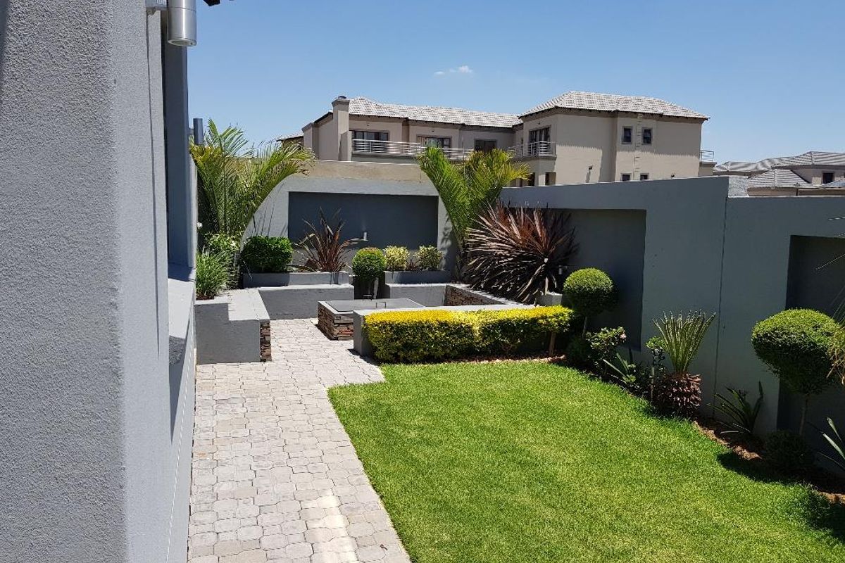 A well-maintained rear garden featuring a paved walkway, manicured lawn, built-in fire pit area, and structured retaining walls.