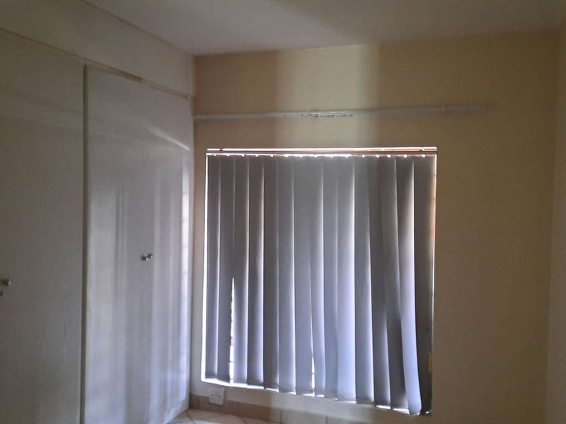 A simple, empty bedroom featuring built-in cupboards, tiled flooring, and vertical blinds over the window.
