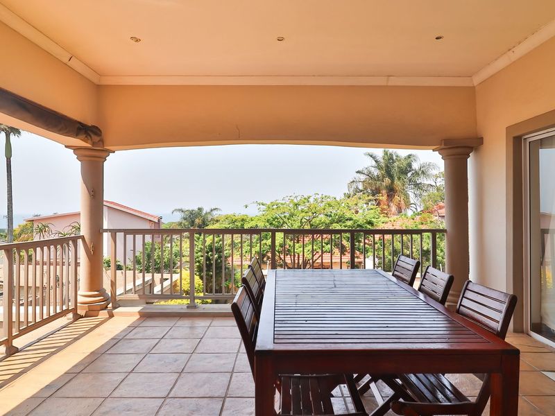 A spacious covered patio featuring tiled flooring, decorative columns, and a metal balustrade, offering an open-air space with views of the surrounding neighborhood.