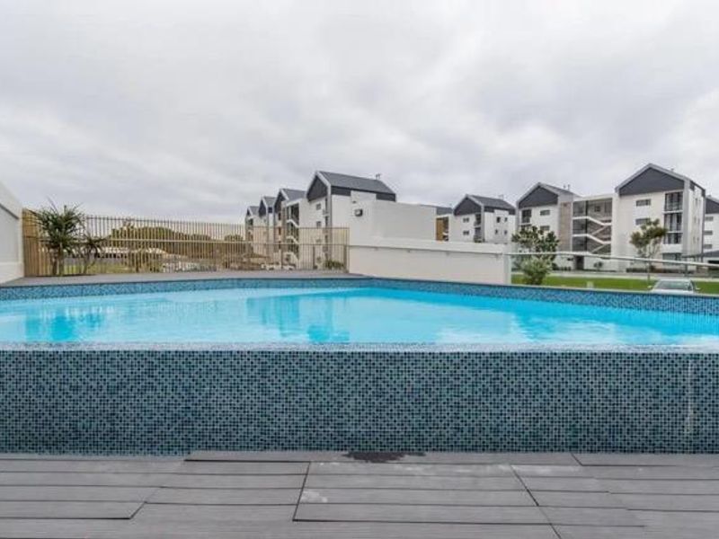 A well-maintained swimming pool area featuring modern blue mosaic tiling, timber decking, and secure metal perimeter fencing within an apartment complex.