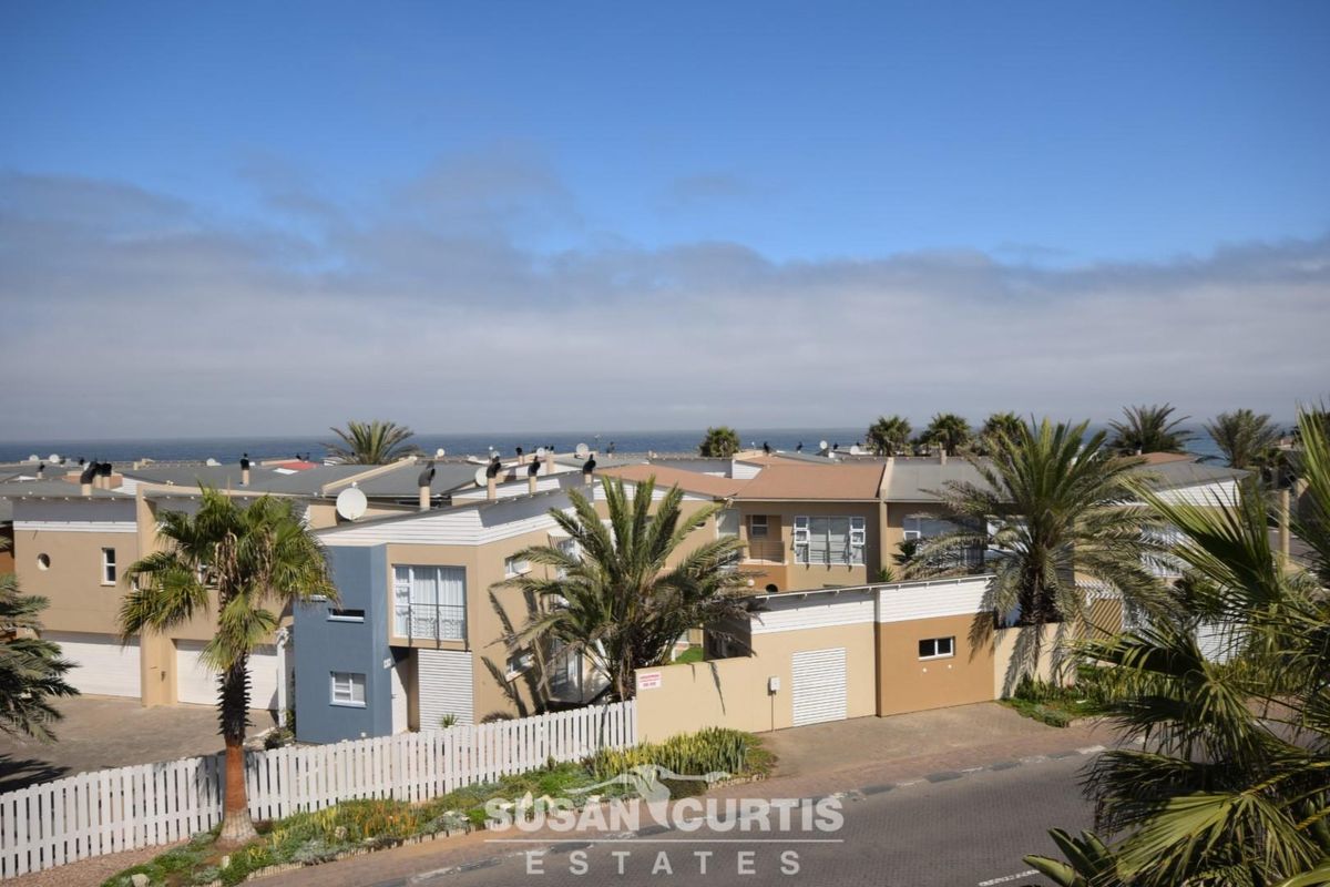 An elevated view of a residential complex featuring modern townhouses with garages, palm trees, and a coastal backdrop.