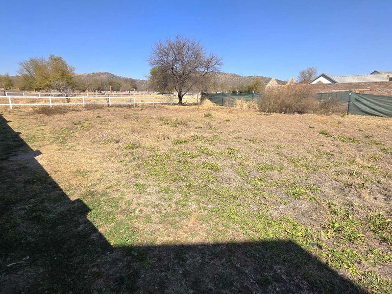A vacant residential plot featuring dry grass, a brick boundary wall, and perimeter fencing with distant mountain views.