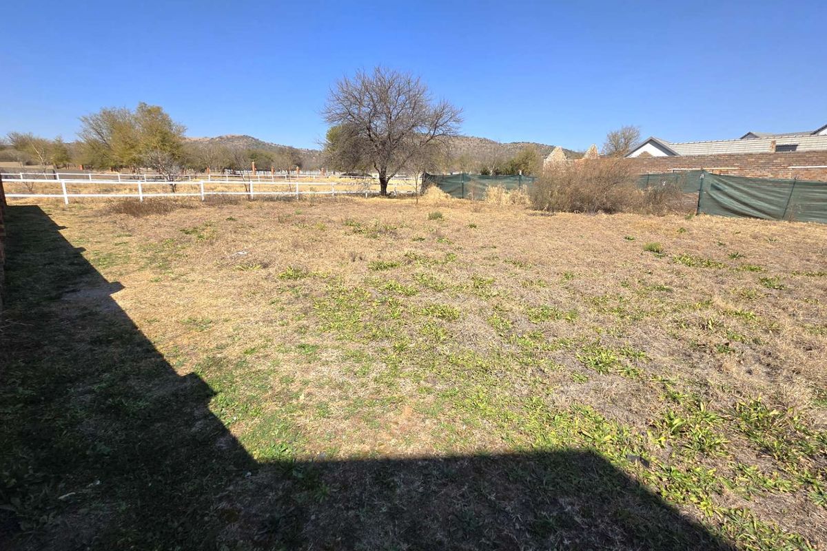 A vacant residential plot featuring dry grass, a brick boundary wall, and perimeter fencing with distant mountain views.