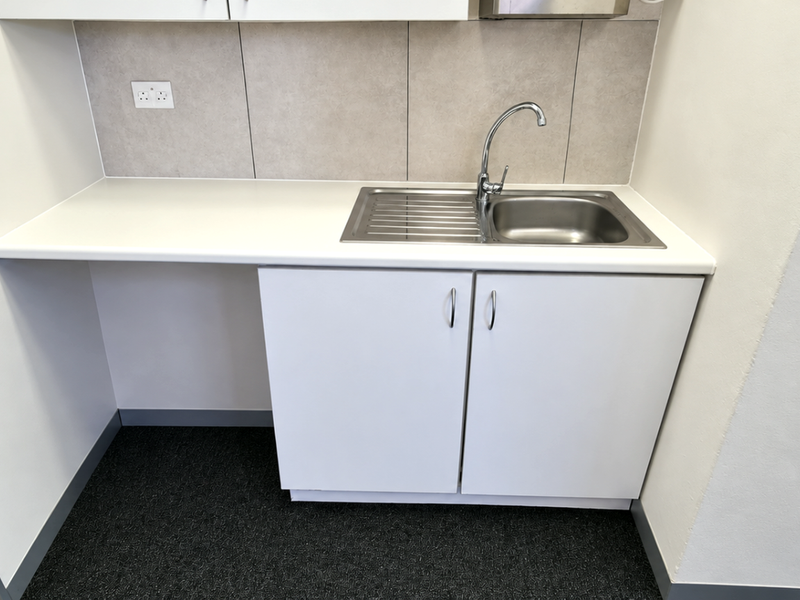 A clean, modern kitchenette area featuring white cabinetry, a stainless steel sink, and a neutral tiled backsplash in an office setting.