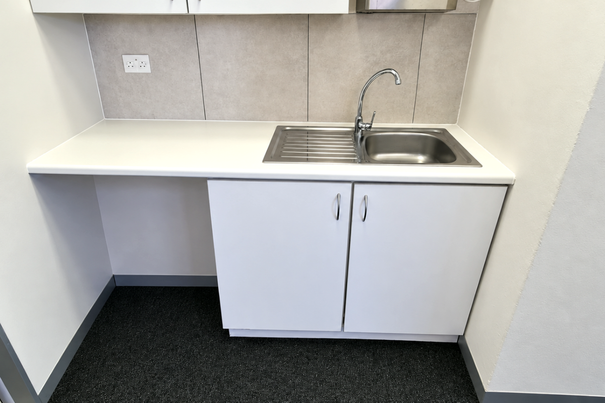 A clean, modern kitchenette area featuring white cabinetry, a stainless steel sink, and a neutral tiled backsplash in an office setting.