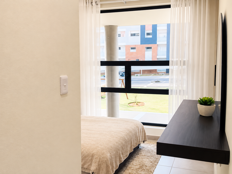 A bright bedroom featuring a large window with natural light, a modern floating desk, and a wall-mounted mirror, overlooking a residential complex.