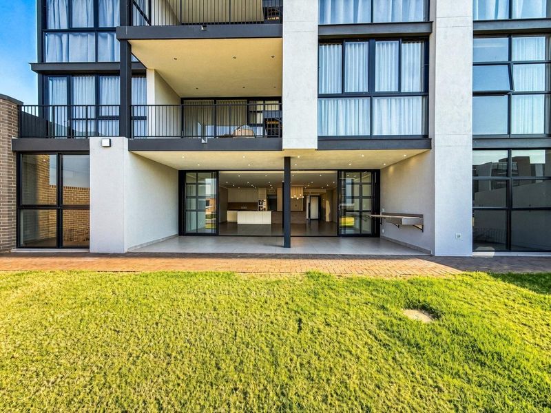 Rear view of a modern apartment building featuring a ground-floor covered patio with sliding doors, brick paving, and an upper-level balcony.