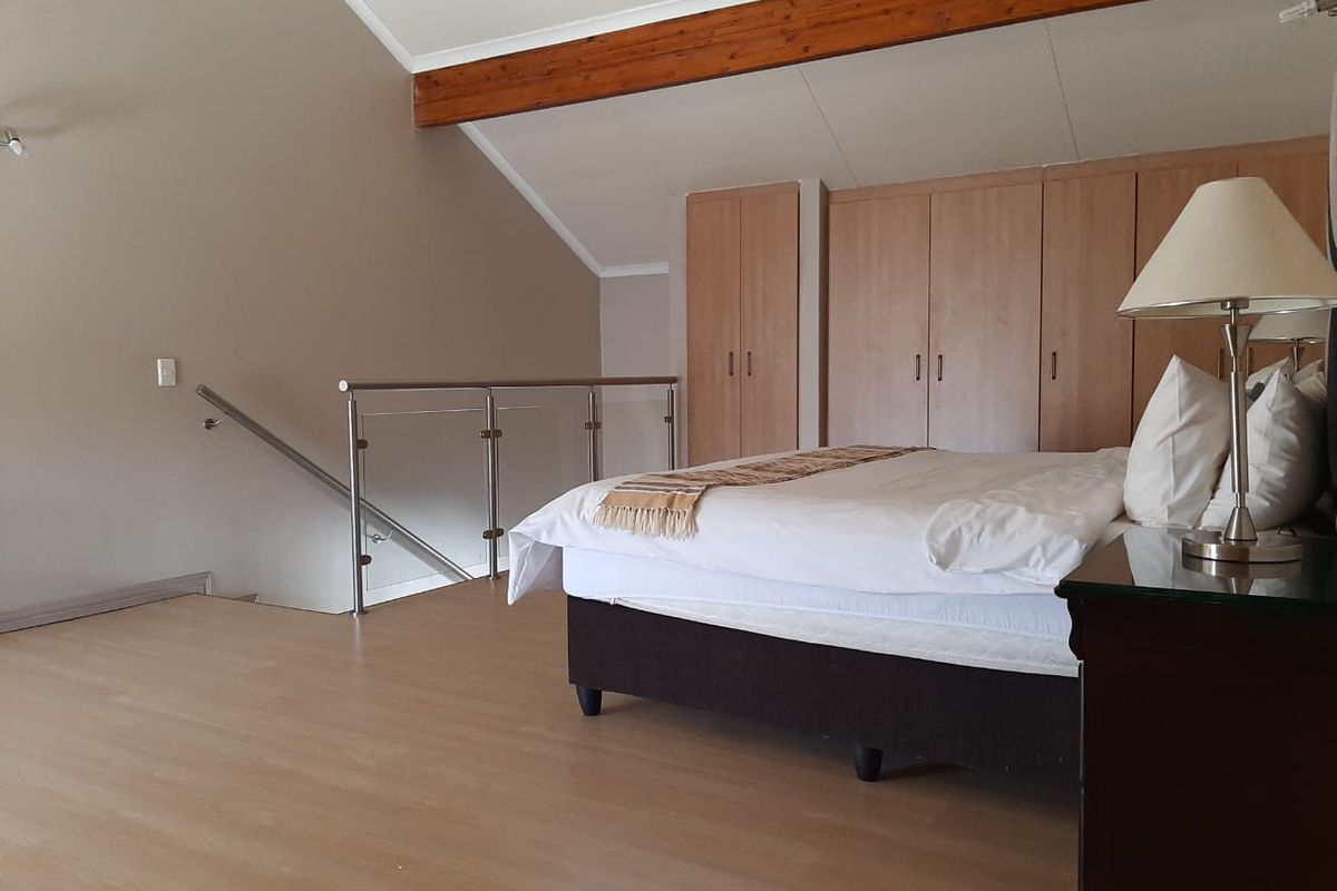 Spacious loft-style bedroom featuring built-in wooden cupboards, exposed timber ceiling beams, and a modern glass balustrade overlooking the lower level.