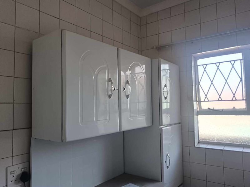 A dated kitchen area featuring white wall-mounted cabinetry and fully tiled walls with a window fitted with security burglar bars.