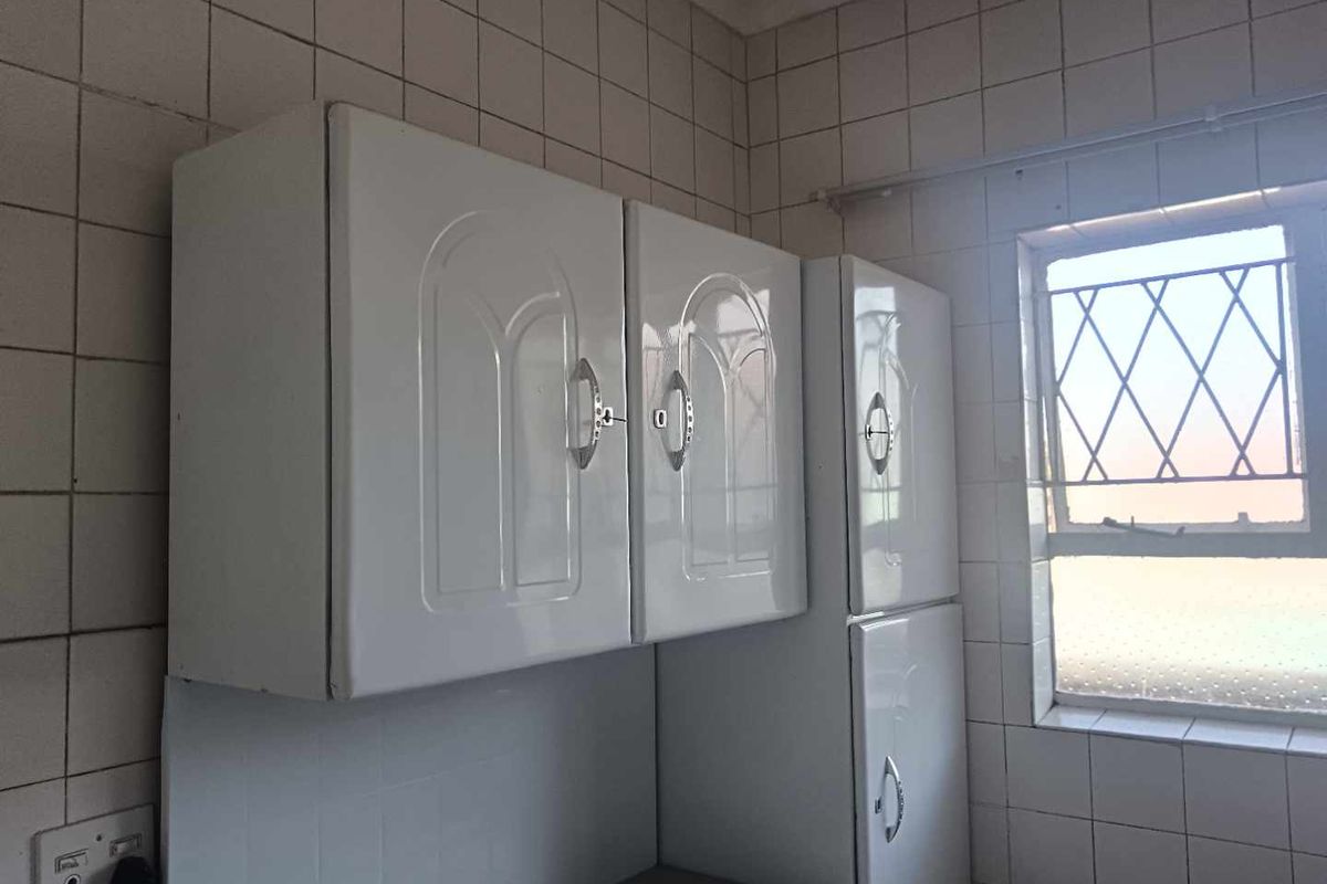 A dated kitchen area featuring white wall-mounted cabinetry and fully tiled walls with a window fitted with security burglar bars.
