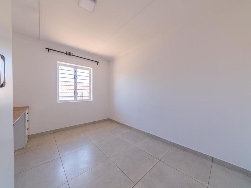 A clean, empty bedroom featuring neutral tiled flooring, a built-in cupboard, and a window fitted with security bars.